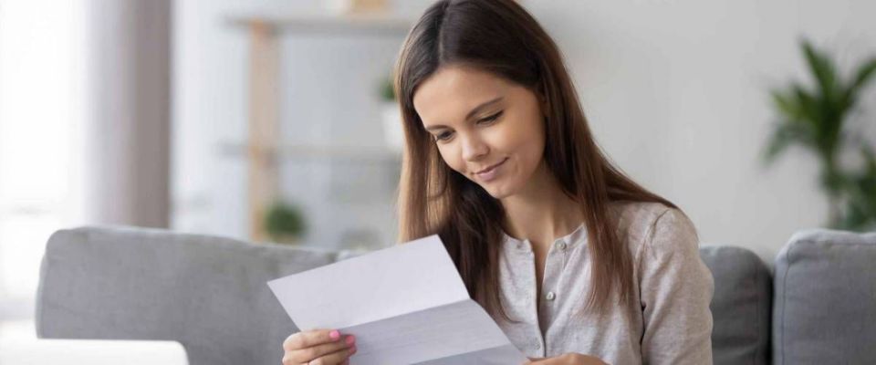 Young woman sitting on sofa at home holds paper reading letter looks happy