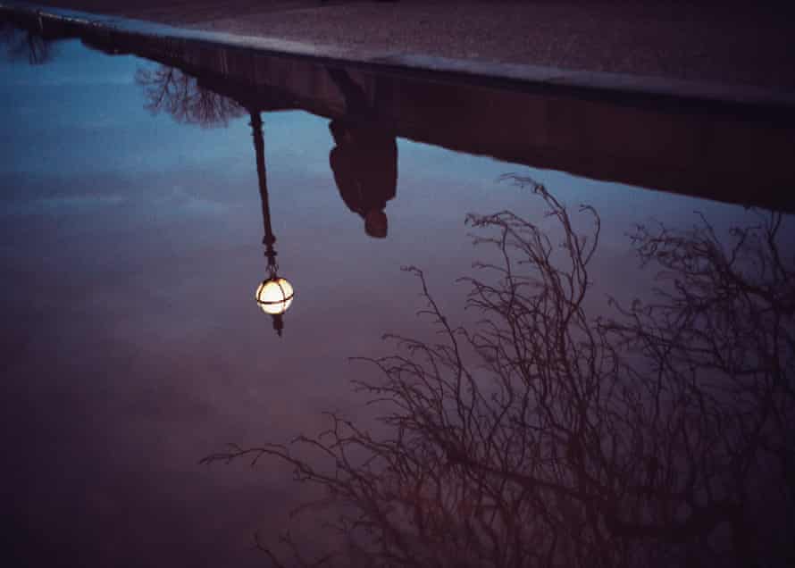 Photo shows a reflection of a figure in a puddle of water while walking a city street late at night.