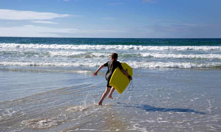 young girl wearing wetsuit running into sea with surfboard