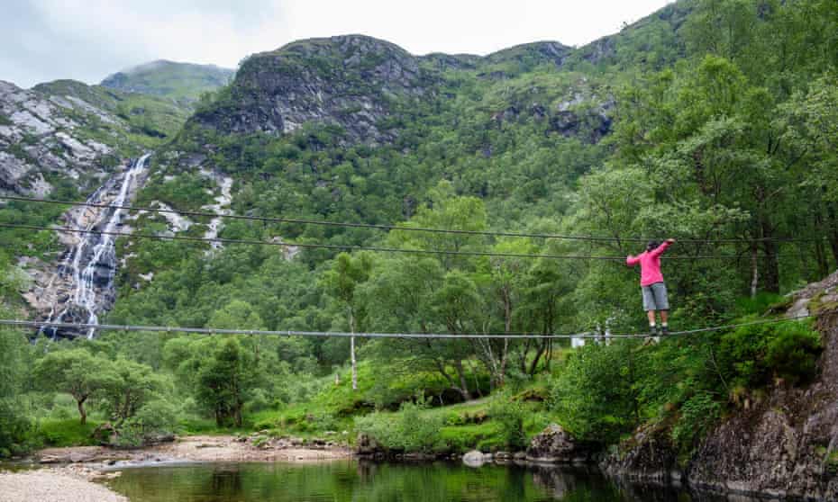 Woman walking across the An Steall wire bridge over the Water of Nevis river with Steall Falls beyond, in Scotland, UK.
