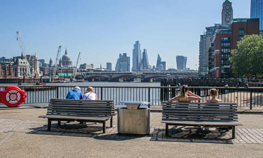 People on benches in the sunshine on London's South Bank looking across to the city skyline and financial district